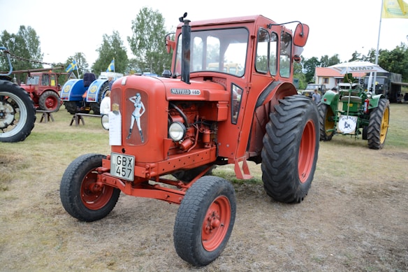 A vintage red tractor with large black tires is prominently displayed on a grassy field. The tractor features a classic design with an illustration of a pin-up girl on its front. Other tractors of various colors and designs can be seen in the background, accompanied by Swedish flags, suggesting an agricultural or vintage vehicle event. Several trees and a small building are visible in the distance.