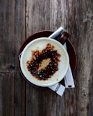 A cup of coffee with frothy milk and a drizzle of caramel sauce on top, set on a brown saucer. Two sugar packets lie beside the cup on a rustic wooden table.