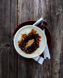 A cup of coffee with frothy milk and a drizzle of caramel sauce on top, set on a brown saucer. Two sugar packets lie beside the cup on a rustic wooden table.