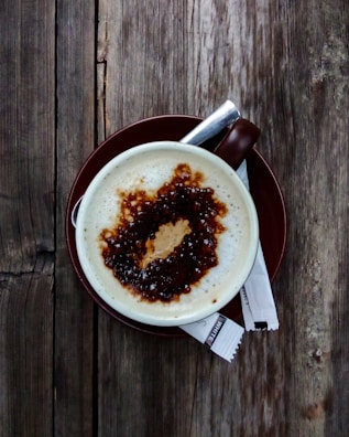 A cup of coffee with frothy milk and a drizzle of caramel sauce on top, set on a brown saucer. Two sugar packets lie beside the cup on a rustic wooden table.