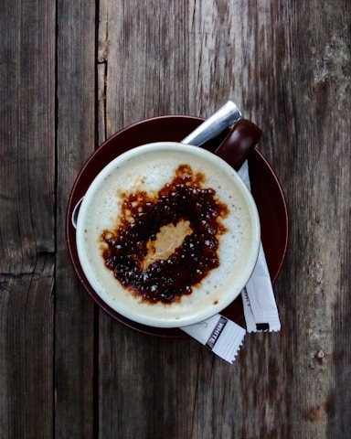 A cup of coffee with frothy milk and a drizzle of caramel sauce on top, set on a brown saucer. Two sugar packets lie beside the cup on a rustic wooden table.