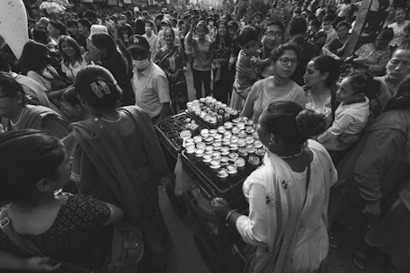 A candid black-and-white photo of a bustling street corner with diverse people interacting under soft afternoon light.