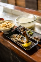 A traditional Japanese meal displayed on a wooden countertop. The meal includes a bowl of rice with vegetables, a glass bowl of soup or broth, and a rectangular plate with grilled fish, lemon wedge, and various garnishes.