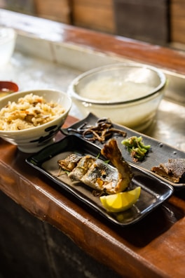 A traditional Japanese meal displayed on a wooden countertop. The meal includes a bowl of rice with vegetables, a glass bowl of soup or broth, and a rectangular plate with grilled fish, lemon wedge, and various garnishes.