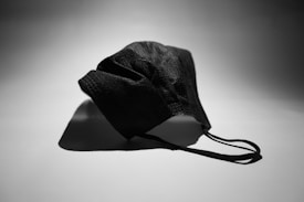 A black face mask with ear loops lies on a flat surface, casting a shadow. The lighting creates a stark contrast between the mask and the background.