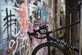 A sleek black bicycle with drop handlebars is positioned against a brick wall covered in colorful and varied graffiti. The bicycle has a label 'AURUM' and a model name 'MAGMA'. The setting appears urban with elements such as a tree and some plants peaking in from the right side.