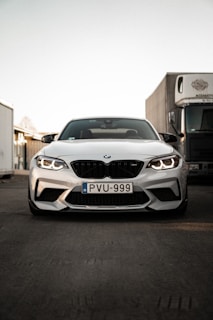 A white BMW car is parked on an industrial street with its headlights on. The car is oriented directly towards the camera, emphasizing its grille and design features. In the background, there are industrial buildings and a large truck, suggesting an urban or industrial setting.