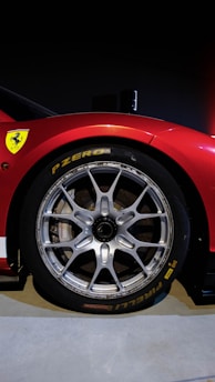 A close-up view of a car wheel featuring a sleek silver alloy rim and a Pirelli tire. The car body is red and bears a yellow logo with a black horse, symbolizing a luxury sports car brand.