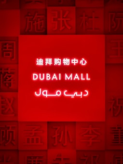 A brightly illuminated red sign featuring the words 'Dubai Mall' in English, Chinese, and Arabic. Surrounding the central text are various Chinese characters embossed into the red background.