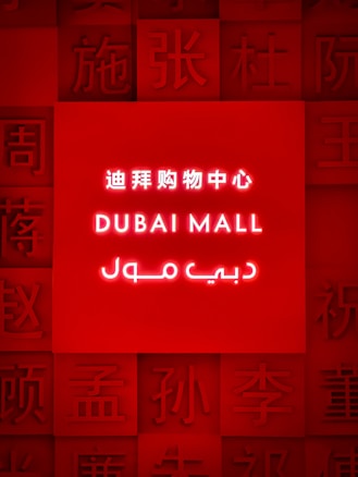 A brightly illuminated red sign featuring the words 'Dubai Mall' in English, Chinese, and Arabic. Surrounding the central text are various Chinese characters embossed into the red background.
