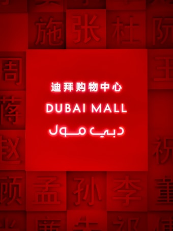 A brightly illuminated red sign featuring the words 'Dubai Mall' in English, Chinese, and Arabic. Surrounding the central text are various Chinese characters embossed into the red background.