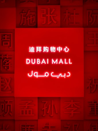 A brightly illuminated red sign featuring the words 'Dubai Mall' in English, Chinese, and Arabic. Surrounding the central text are various Chinese characters embossed into the red background.