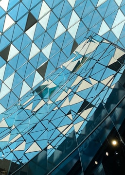 A geometric pattern of triangular glass panels, creating a reflective surface. The panels form a grid-like structure with shades of blue and white.