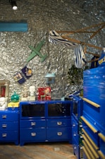 A creatively styled room features bright blue cabinets with metallic handles, topped with various decorative objects such as a green glass orb, a white bust, and small figurines. The walls are covered with silver foil, giving a futuristic and industrial feel. A green wooden airplane, a striped hammock, and a blue backpack are suspended for added visual interest.