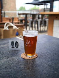 A pint of amber-colored beer rests on a round coaster on a textured table. The setting appears to be outdoors, with blurred wooden tables and metal stools in the background. A small table number sign displaying the number 10 is placed next to the glass, and a bag with a strap and a blue tassel sits in the background.