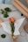 A single orange and pink rose is wrapped in brown paper with a white tissue inside. The rose is placed on a table alongside green leaves, a roll of brown paper, and a spool of twine. The setting has a crafting theme with a simple, natural aesthetic.
