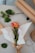 A single orange and pink rose is wrapped in brown paper with a white tissue inside. The rose is placed on a table alongside green leaves, a roll of brown paper, and a spool of twine. The setting has a crafting theme with a simple, natural aesthetic.