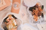 Two containers hold desserts that are creatively arranged with crispy rolls, pretzels, and cream, decorated with small garnishes. The background contains packaging and other elements, including paper designed to look like newsprint.