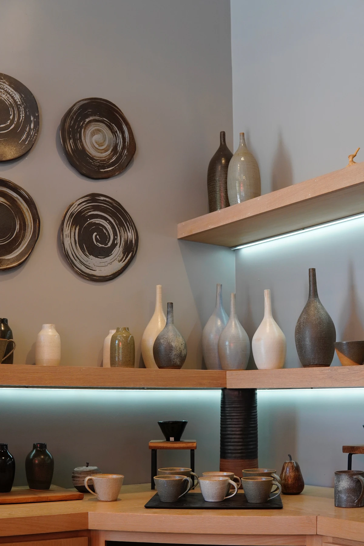 A quiet, earthy room corner featuring open wooden shelves filled with mismatched raw clay vases, stone mugs, and simple neutral decorations.