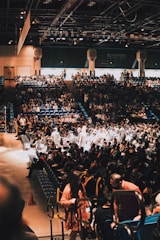 A large indoor auditorium filled with many people, including rows of seated individuals wearing graduation caps and gowns. Some attendees are wearing casual attire. The environment is bustling, with the audience sitting in tiered seating while focusing on the stage area where graduates are gathered.