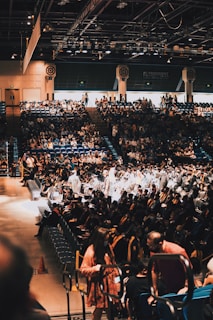 A large indoor auditorium filled with many people, including rows of seated individuals wearing graduation caps and gowns. Some attendees are wearing casual attire. The environment is bustling, with the audience sitting in tiered seating while focusing on the stage area where graduates are gathered.