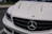The image features the front portion of a white luxury car, prominently displaying the emblem on the hood. The sleek design includes distinctive headlight styling and a black grille adding contrast to the white exterior.