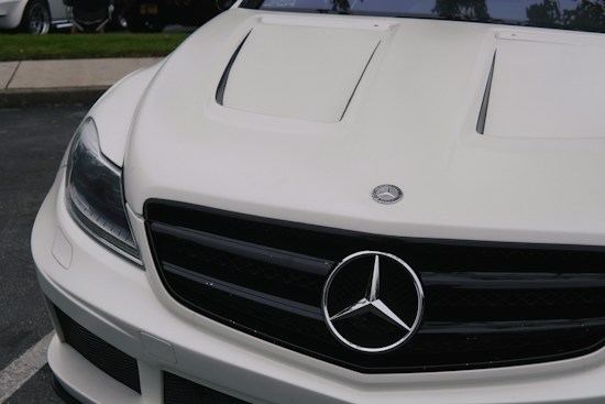 The image features the front portion of a white luxury car, prominently displaying the emblem on the hood. The sleek design includes distinctive headlight styling and a black grille adding contrast to the white exterior.