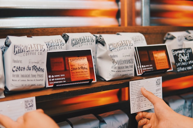 Several neatly arranged bags of coffee from Chromatic Coffee Co. are displayed on a wooden shelf. Each bag has detailed labeling with names like 'Côtes du Rhône' and 'Bourbogne,' suggesting different blends or origins. A hand is holding a card with additional information about the coffee.