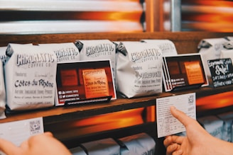 Several neatly arranged bags of coffee from Chromatic Coffee Co. are displayed on a wooden shelf. Each bag has detailed labeling with names like 'Côtes du Rhône' and 'Bourbogne,' suggesting different blends or origins. A hand is holding a card with additional information about the coffee.