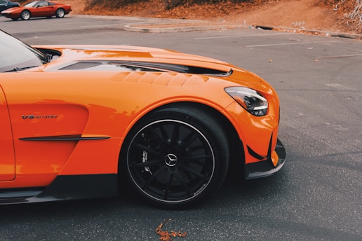 A sleek orange sports car with a V8 biturbo engine badge is parked on an asphalt surface. The car features black wheels and aerodynamic body styling. In the background, another red car is partially visible, with autumn leaves scattered around.