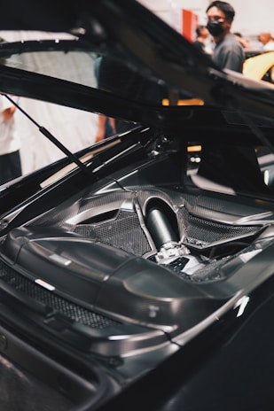 A detailed view of a high-performance car engine with a sleek carbon fiber finish. The engine is displayed prominently under an open hood, while several people are visible in the background, some wearing face masks.