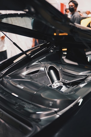 A detailed view of a high-performance car engine with a sleek carbon fiber finish. The engine is displayed prominently under an open hood, while several people are visible in the background, some wearing face masks.