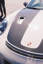 A close-up view of the front of a sports car displaying a carbon fiber hood with aerodynamic vents and a prominent logo crest. The car has a sleek, polished finish, and the background shows an empty parking area. Part of a person’s legs in black pants and casual shoes are visible on the left side, standing near the car.