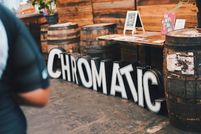 Large black and white letters spelling 'CHROMATIC' are arranged on the floor beside a row of wooden barrels. In the background, a wooden table displays various colorful posters and flyers, with additional chalkboard signs and floral arrangements.