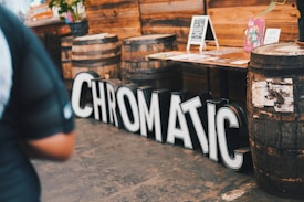 Large black and white letters spelling 'CHROMATIC' are arranged on the floor beside a row of wooden barrels. In the background, a wooden table displays various colorful posters and flyers, with additional chalkboard signs and floral arrangements.
