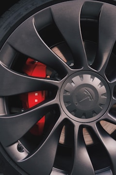 A close-up view of a matte black car wheel rim featuring a series of sleek, aerodynamic spokes with a central emblem. Bright red brake calipers are visible behind the spokes, contributing a bold contrast to the rim's dark color.