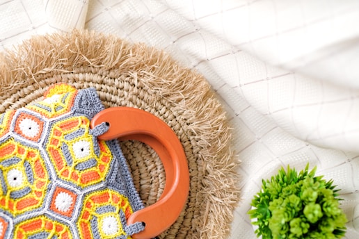 A colorful crocheted bag with geometric patterns in orange, yellow, and grey is placed on a woven straw mat. The bag has a large orange handle, and there is a small green potted plant nearby. The background features a light fabric with subtle grid patterns.