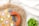 A colorful crocheted bag with geometric patterns in orange, yellow, and grey is placed on a woven straw mat. The bag has a large orange handle, and there is a small green potted plant nearby. The background features a light fabric with subtle grid patterns.