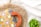 A colorful crocheted bag with geometric patterns in orange, yellow, and grey is placed on a woven straw mat. The bag has a large orange handle, and there is a small green potted plant nearby. The background features a light fabric with subtle grid patterns.