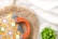 A colorful crocheted bag with geometric patterns in orange, yellow, and grey is placed on a woven straw mat. The bag has a large orange handle, and there is a small green potted plant nearby. The background features a light fabric with subtle grid patterns.