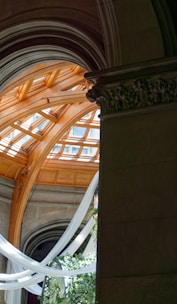 A grand architectural structure featuring intricate stone archways with decorative carvings and a vast, curved wooden ceiling with large windows that allow daylight to stream through. White fabric drapes hang gracefully in the space, complementing the green foliage and white floral arrangements situated below.