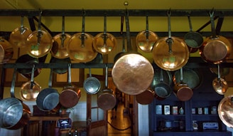 A collection of copper pots and pans hanging from a metal rack in a kitchen setting. The background includes wooden cabinets and a shelf with various kitchen items. The lighting creates a warm and rustic atmosphere.