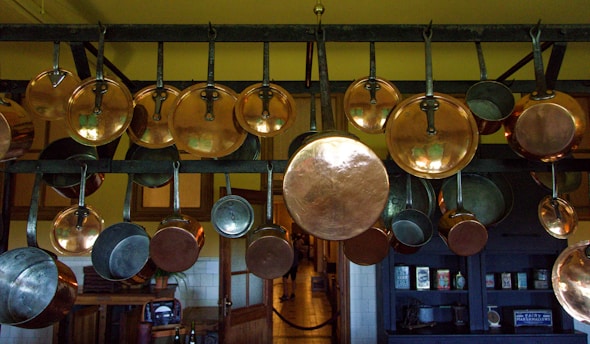 A collection of copper pots and pans hanging from a metal rack in a kitchen setting. The background includes wooden cabinets and a shelf with various kitchen items. The lighting creates a warm and rustic atmosphere.