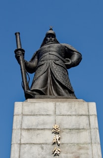 An imposing statue of a warrior in traditional armor stands atop a stone pedestal. The warrior holds a long sword, exuding strength and authority. The base of the statue features inscriptions in an Asian script. The background is a clear, bright blue sky.