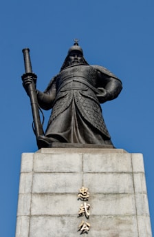 An imposing statue of a warrior in traditional armor stands atop a stone pedestal. The warrior holds a long sword, exuding strength and authority. The base of the statue features inscriptions in an Asian script. The background is a clear, bright blue sky.