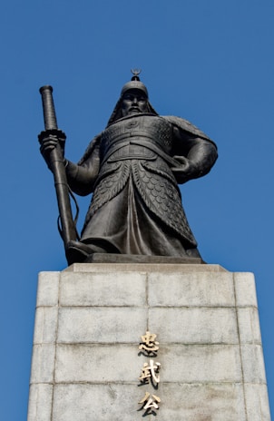 An imposing statue of a warrior in traditional armor stands atop a stone pedestal. The warrior holds a long sword, exuding strength and authority. The base of the statue features inscriptions in an Asian script. The background is a clear, bright blue sky.
