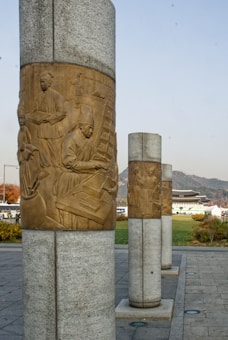 Tall stone columns feature intricate bronze plaques depicting historical figures engaged in scholarly activities. The columns are part of an outdoor setting with a traditional building and mountains visible in the background.