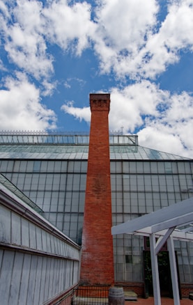 A tall red brick chimney stands prominently against a backdrop of a glass greenhouse structure. The sky is bright with scattered white clouds, providing a stark contrast to the chimney's rugged, industrial appearance. Foreground elements include metal and glass panels along the sides, adding to the geometric aesthetic.