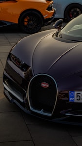 The image features the front part of a luxury sports car with a sleek, aerodynamic design. The logo is prominently displayed on the grille. In the background, parts of two other sports cars can be seen, highlighting a high-performance automotive theme.