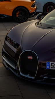 The image features the front part of a luxury sports car with a sleek, aerodynamic design. The logo is prominently displayed on the grille. In the background, parts of two other sports cars can be seen, highlighting a high-performance automotive theme.